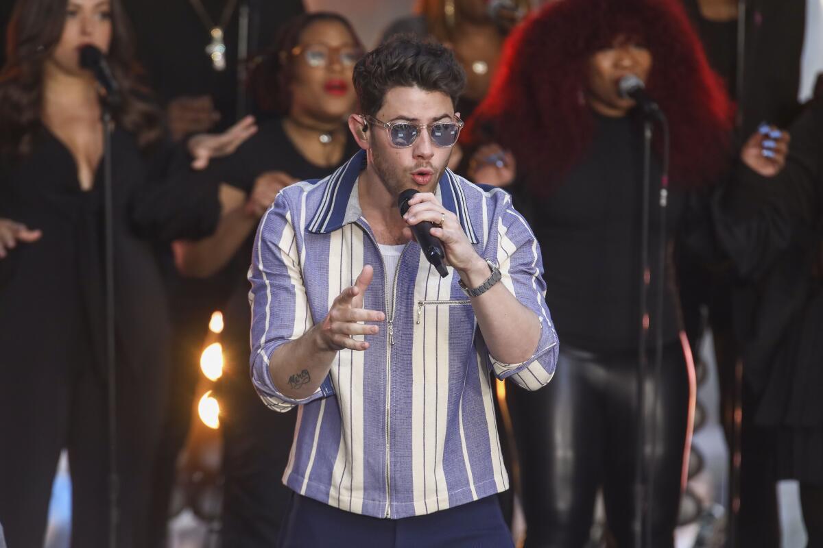 Nick Jonas wears a white-and-blue striped shirt and sunglasses while singing into a microphone.