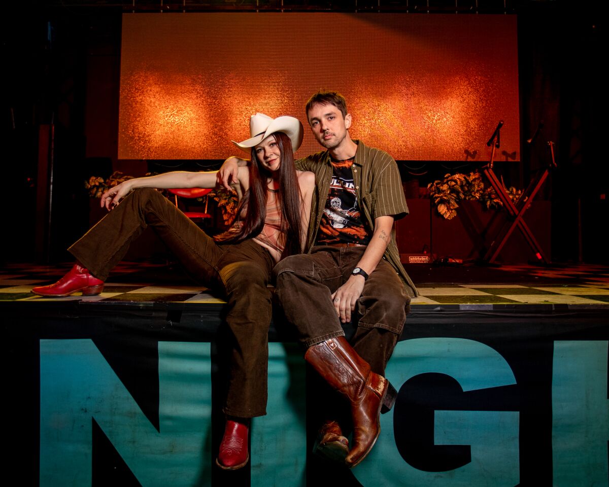A woman in red boots and a cowboy hat sits on a stage with a man in brown cowboy boots
