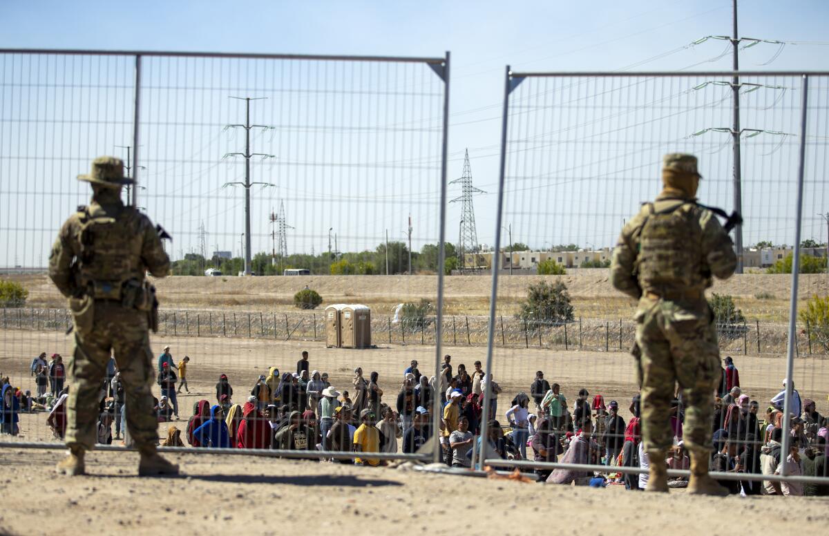 Migrants wait near the border fence as Texas National Guard members watch in El Paso.