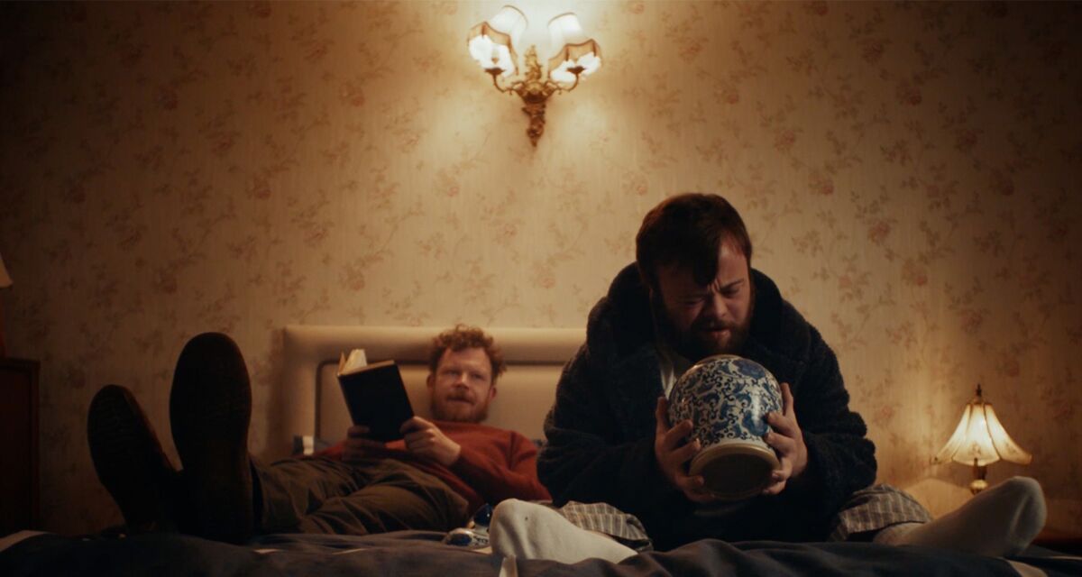 Two men sit on a bed, one holding a book, the other an urn.