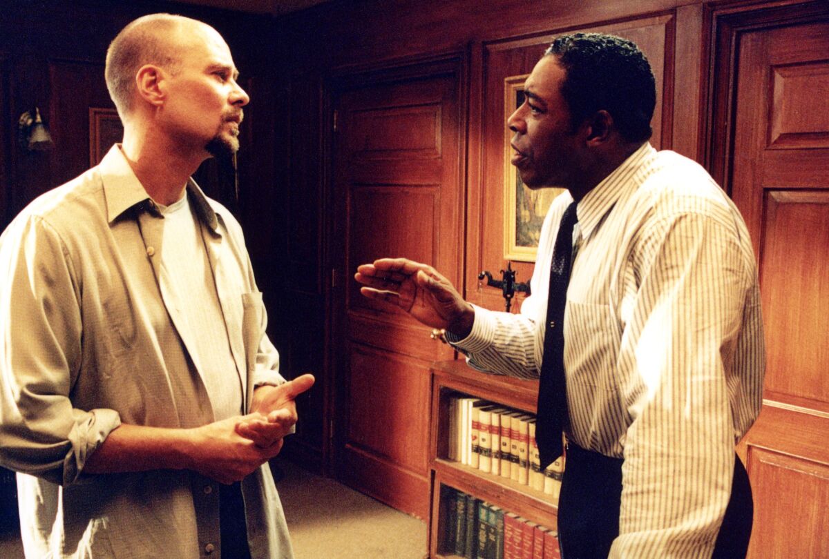 A man in a button-down shirt and tie speaks talks to another man in an office.