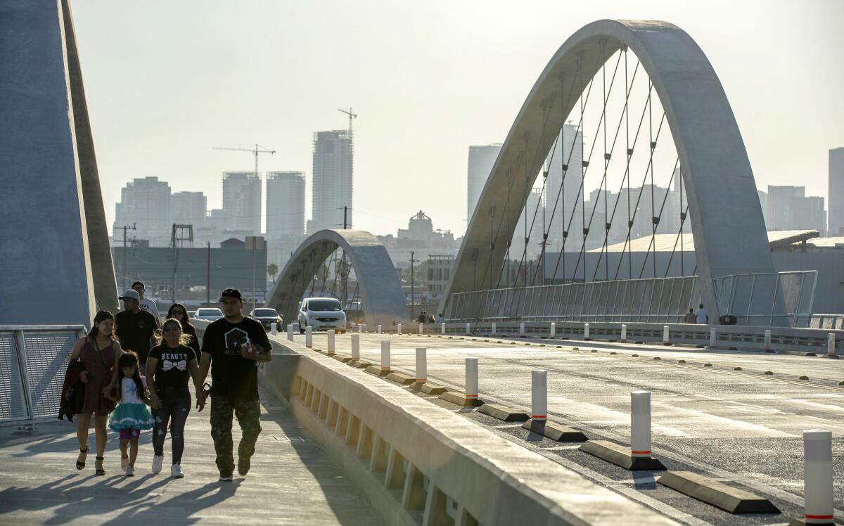 People walk on a bridge with large arches and downtown Los Angeles in the background