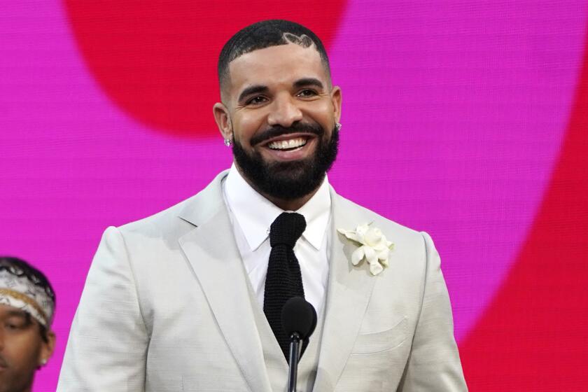 Drake smiles in front of a microphone while wearing a light gray suit and black tie.