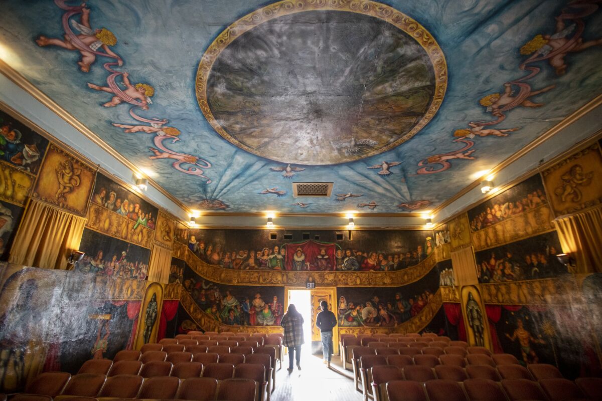 People inside an elaborately decorated building. An audience is painted on the walls; the ceiling looks like a blue sky.