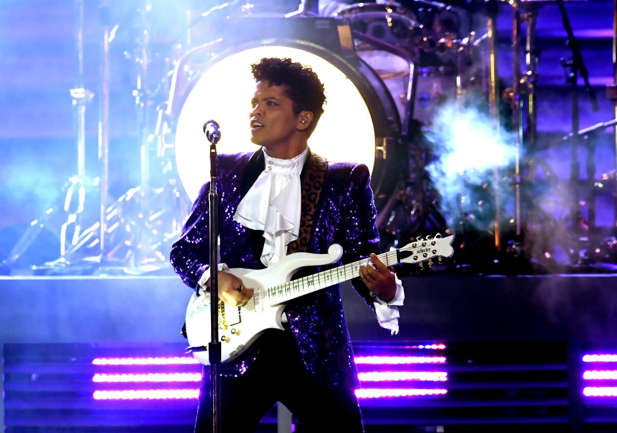 Bruno Mars, in a purple sequined suit, plays guitar and sings on stage.