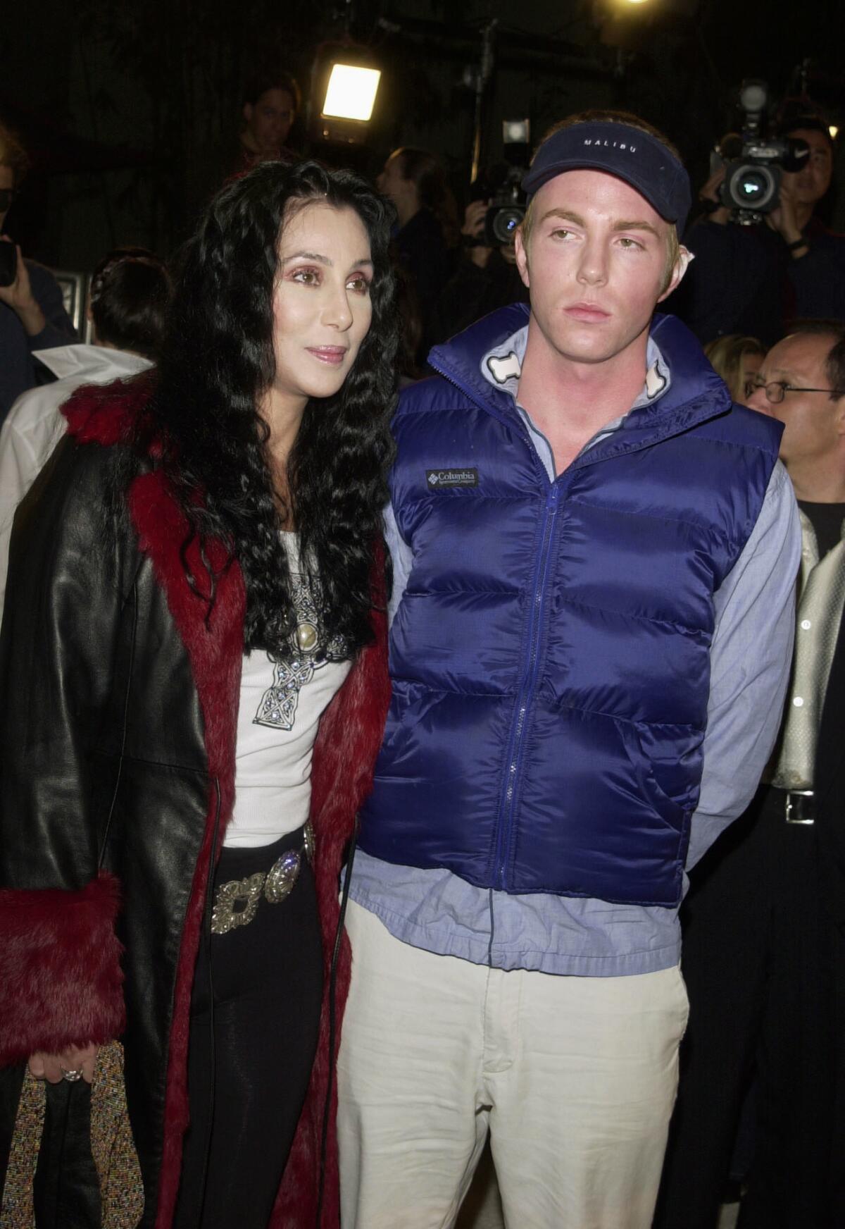 Cher in a black leather trench coat and Elijah Blue Allman in a blue puffer vest stand together at an event