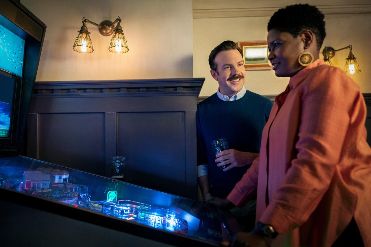 A woman playing pinball while a smiling, mustachioed man looks on.