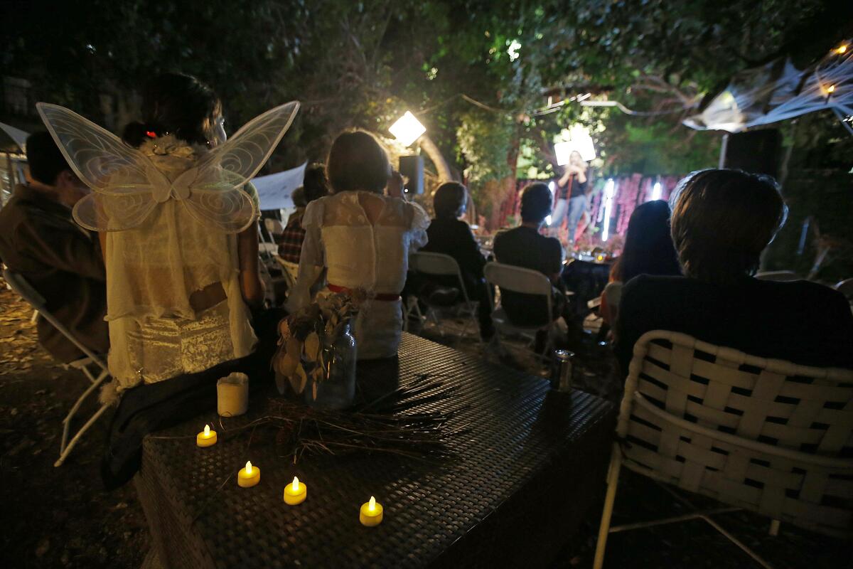 People stand and sit on folding chairs in front of a makeshift stage in a backyard.