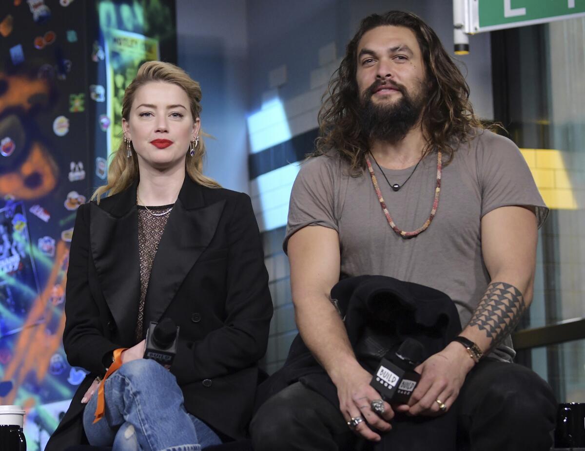 A blond woman and a large bearded man hold microphones while sitting and waiting to answer questions.