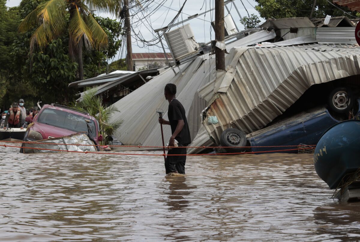 A person walks through a street flooded to his knees. Near him are overturned cars and a caved-in metal roof.