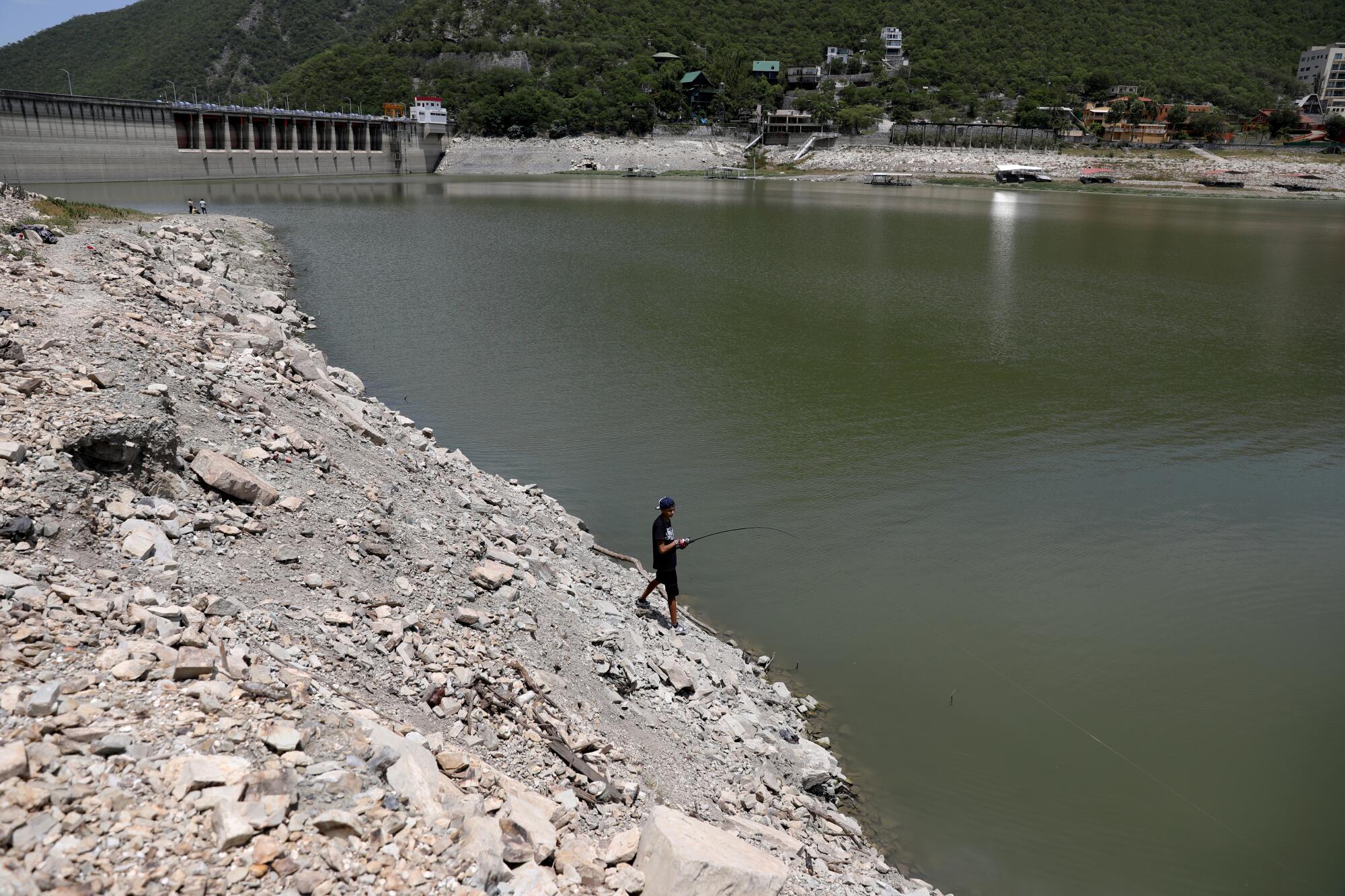 A  boy fishes in a low reservoir with a dam in the distance
