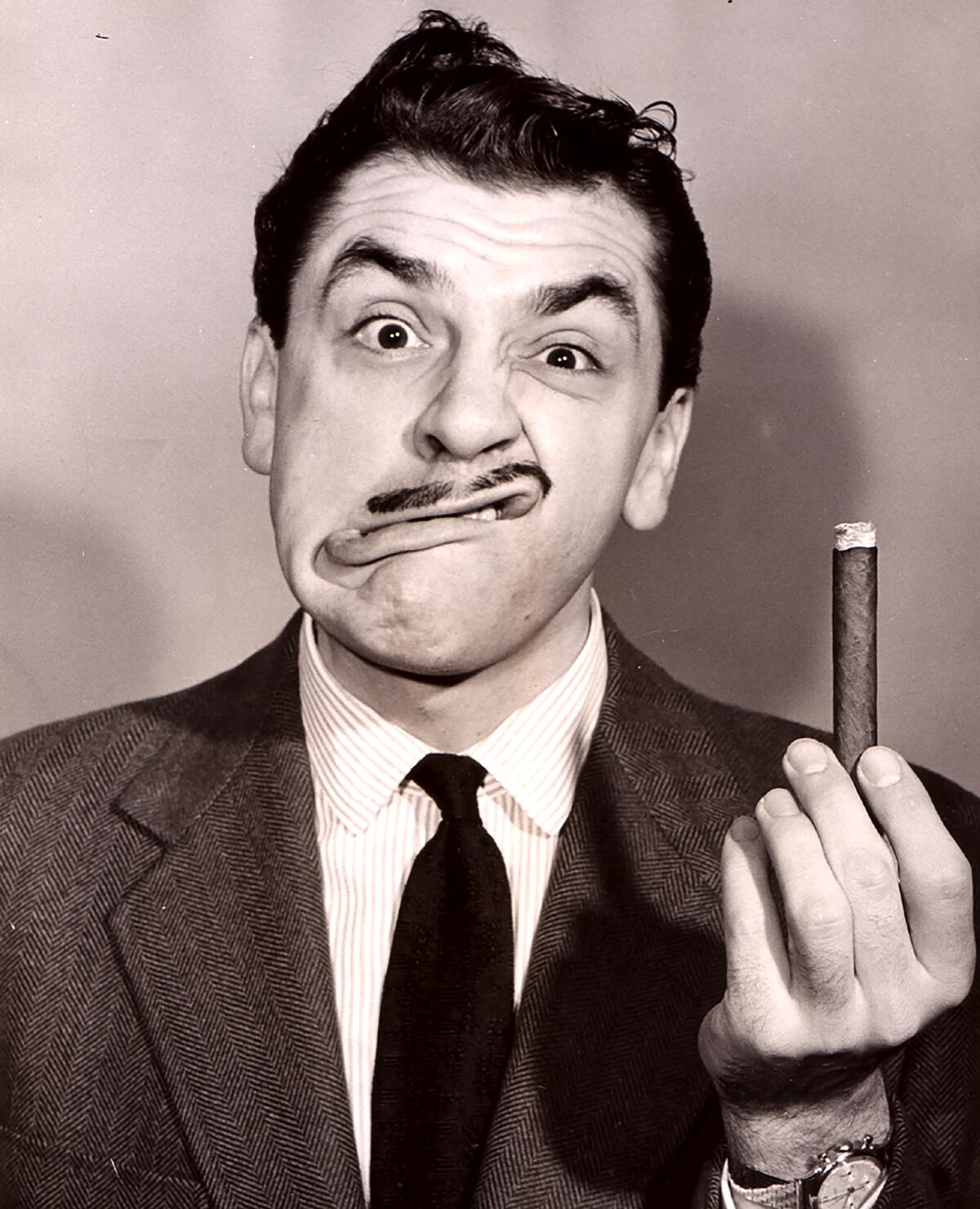 Ernie Kovacs, with his lips askew, holds a cigar straight up in his left hand.