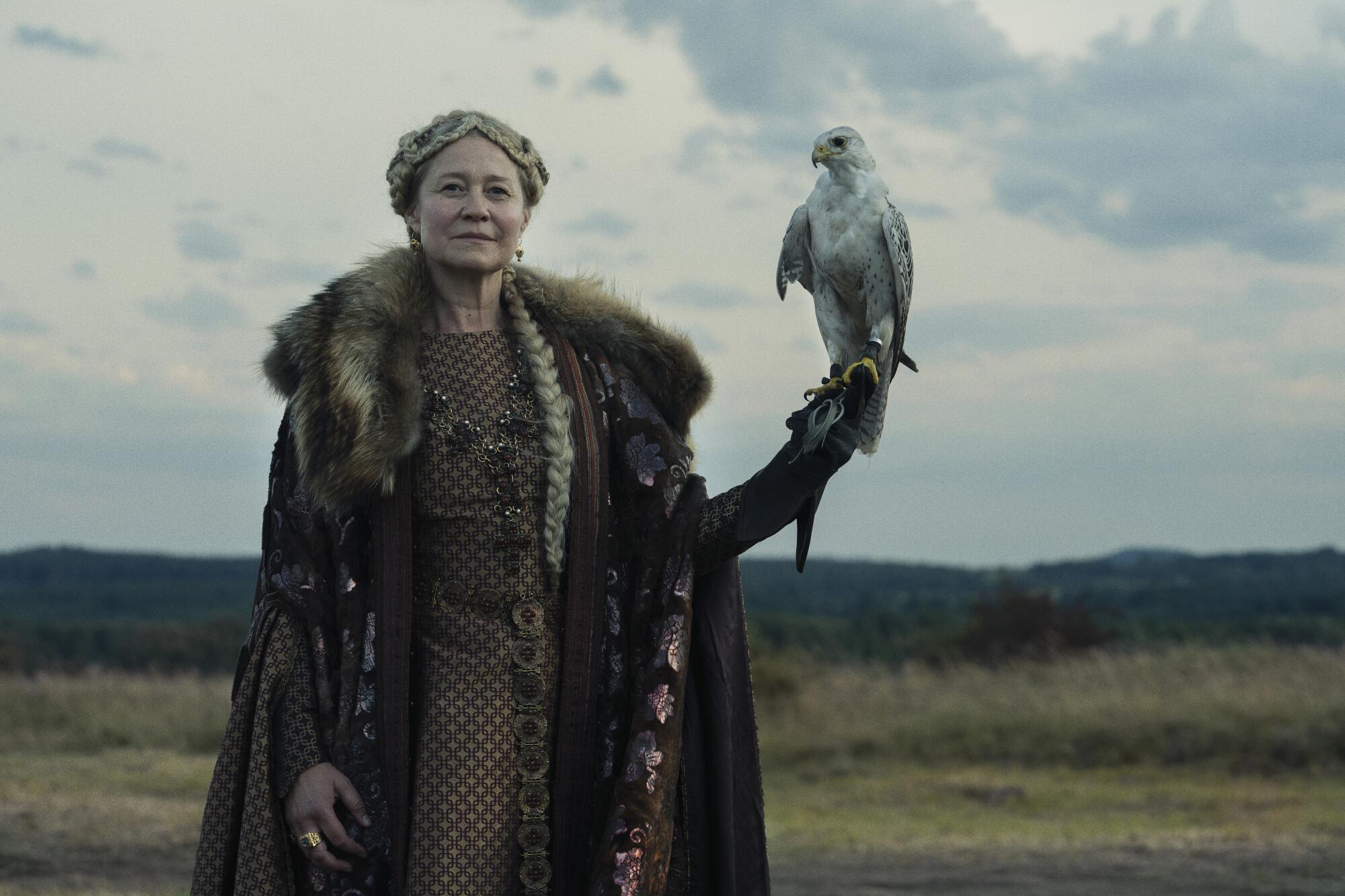 A woman in a fur-collared cape stands outdoors holding a bird of prey on her hand.