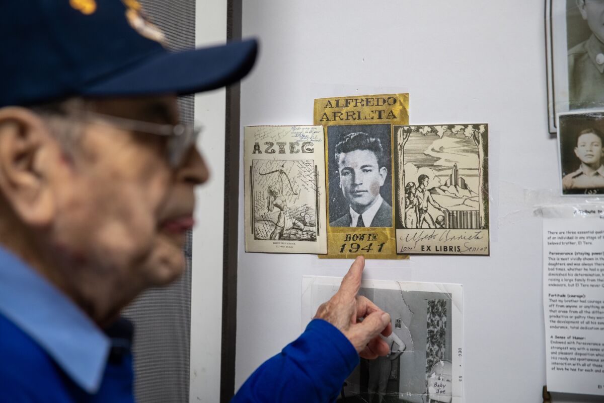 An older man in a baseball cap points to an old photo of himself displayed in his home.