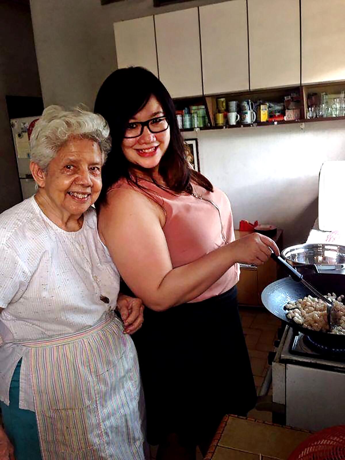 A woman standing by a stove with her grandmother, smiling.