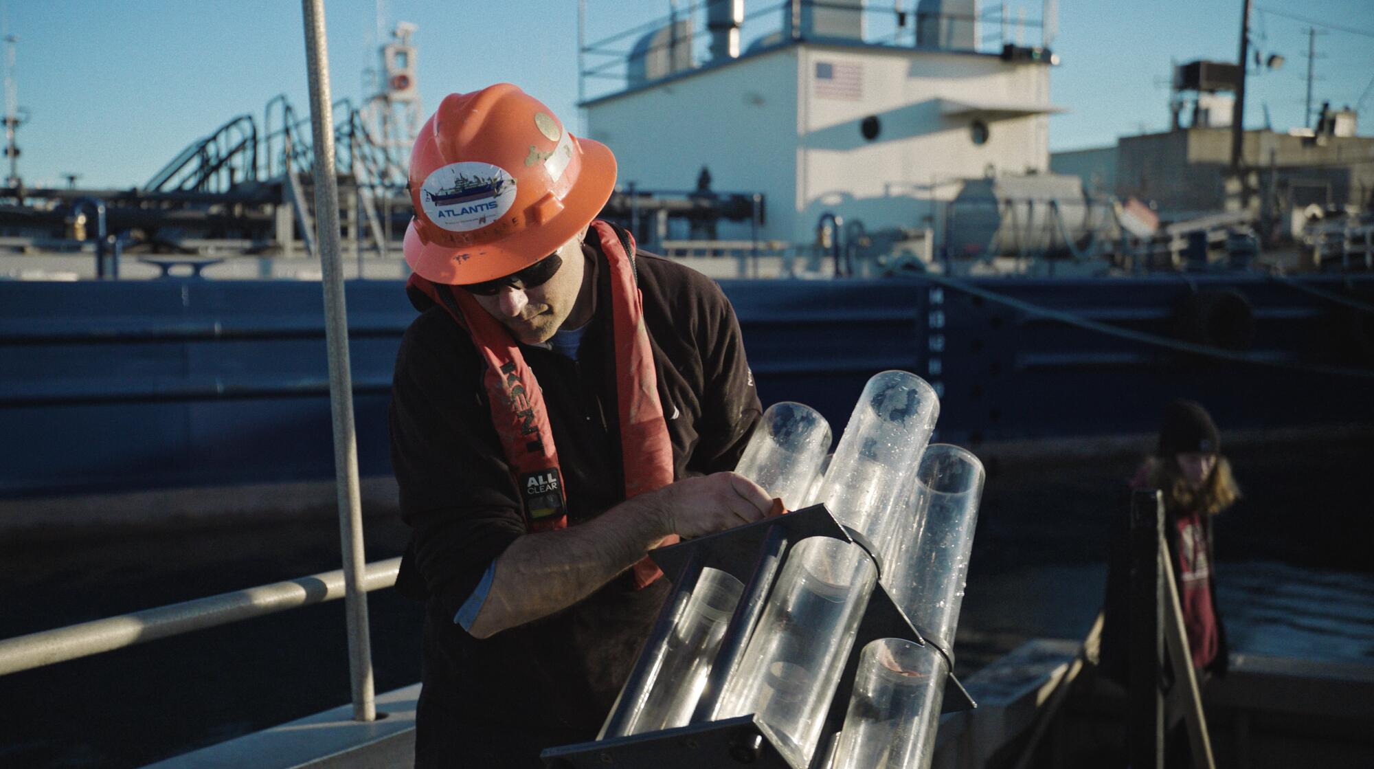 A man wearing an orange hard hat and life vest holds a device made of clear tubes while standing on the deck of a ship.