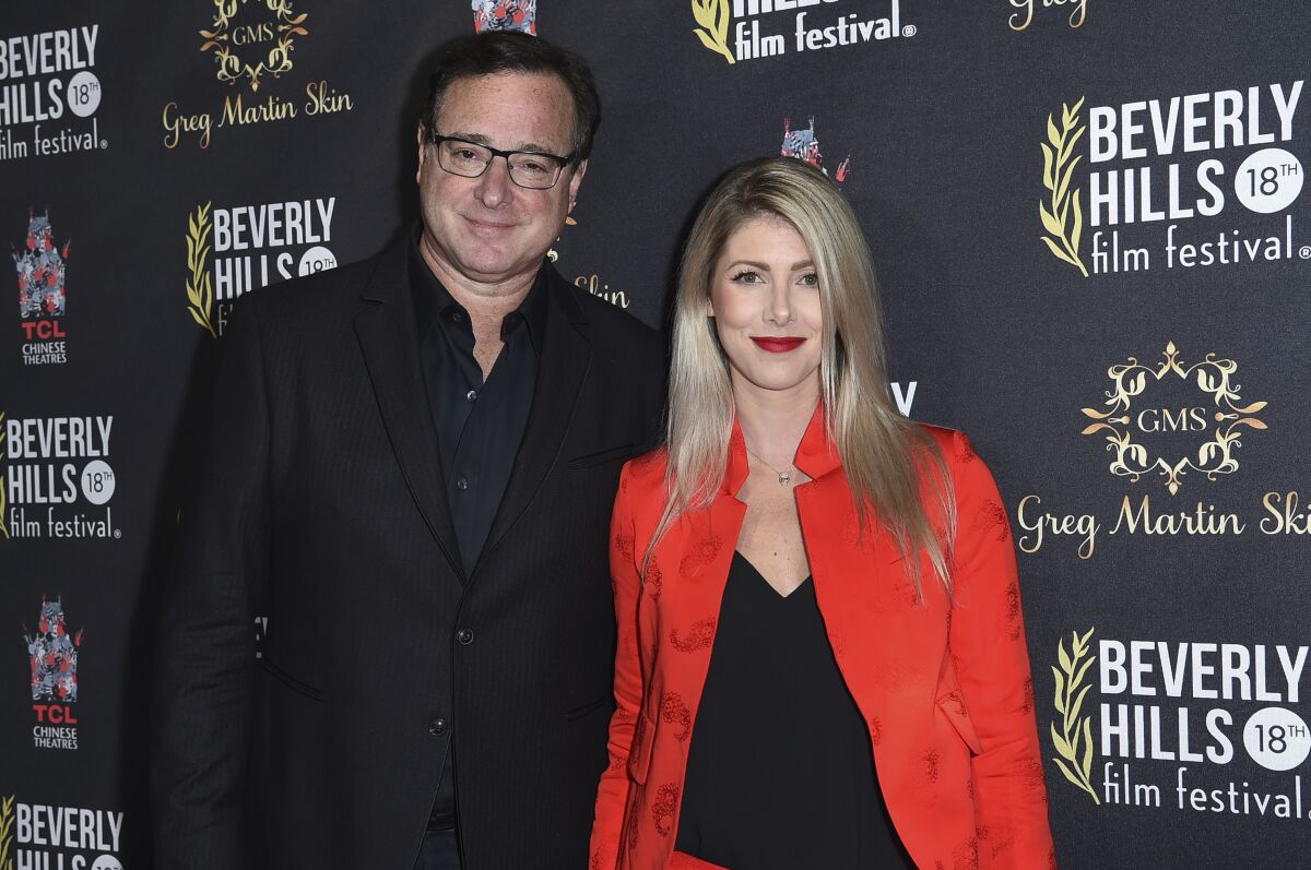 Bob Saget in a black suit posing with Kelly Rizzo in a red jacket