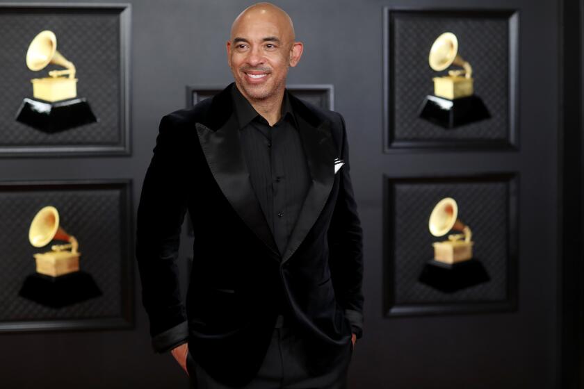 A man in a black tuxedo stands in front of a black wall that has pictures of Grammys on it