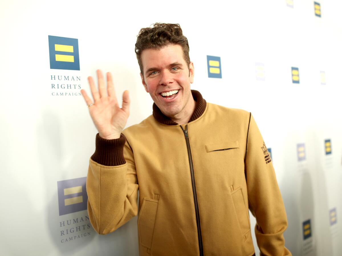 A man in a tan jacket smiles and waves at the camera.