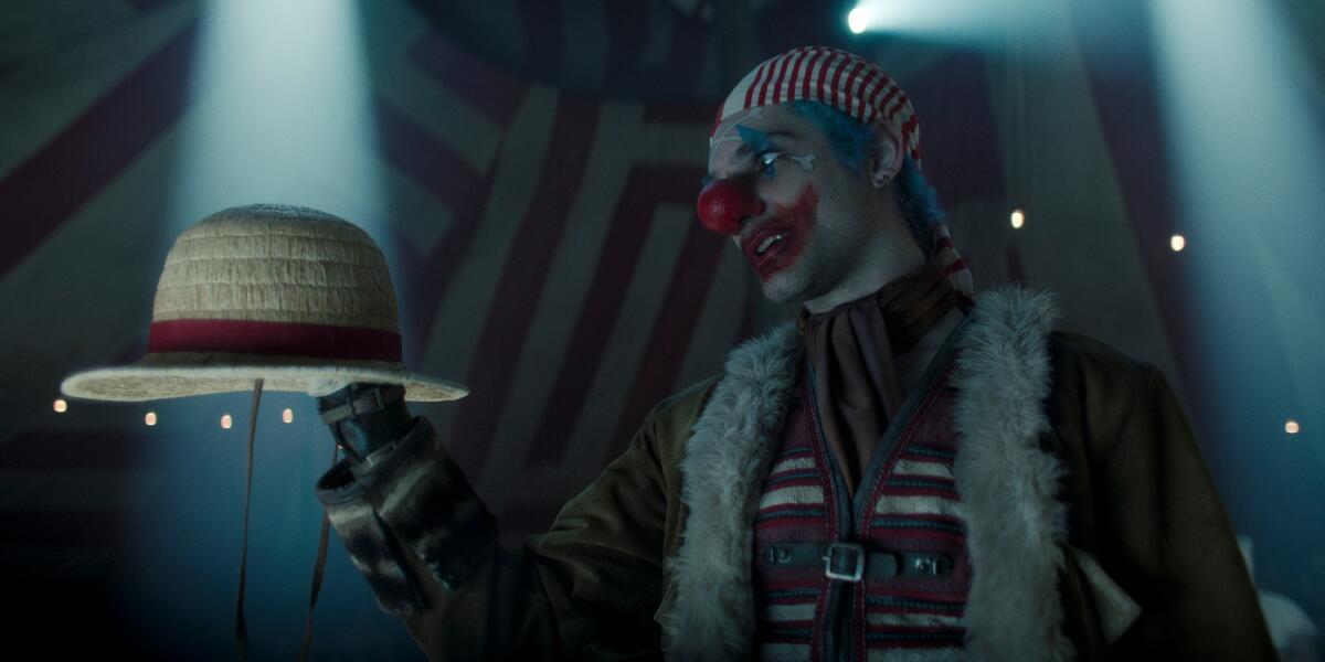 A man in smeared clown makeup, a furled jacket and clown vest holding and looking at a straw hat with a red band