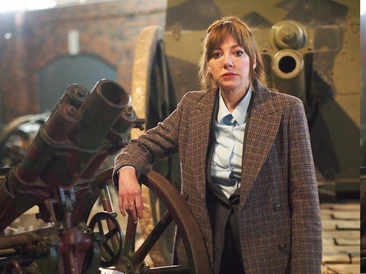 A woman in tweed leaning on an old cannon looking straight into camera.