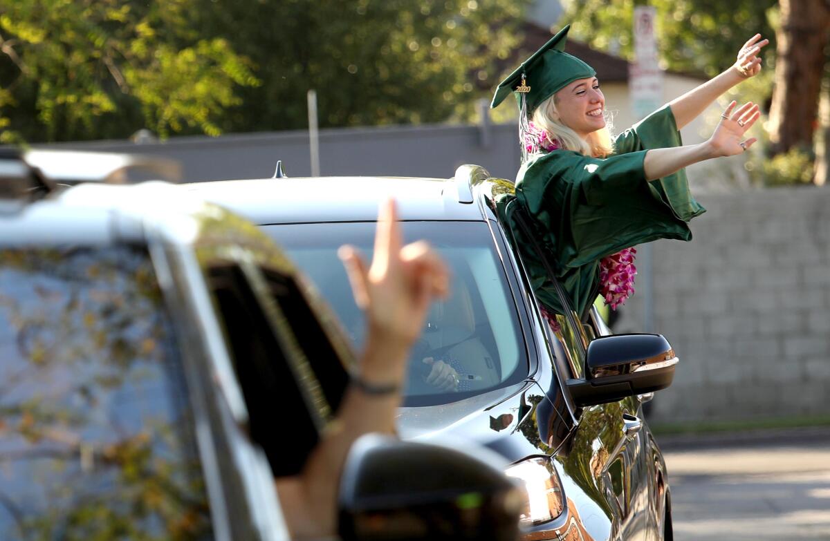 A high school graduate in green cap and gown waves to her teachers through the open window of a car