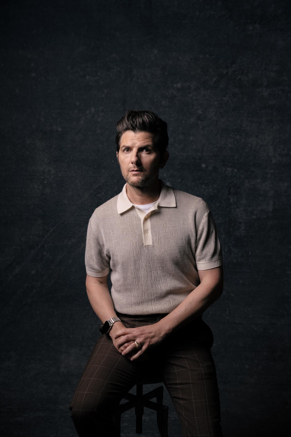 Actor Adam Scott sits on a stool in front of a dark background for a portrait.