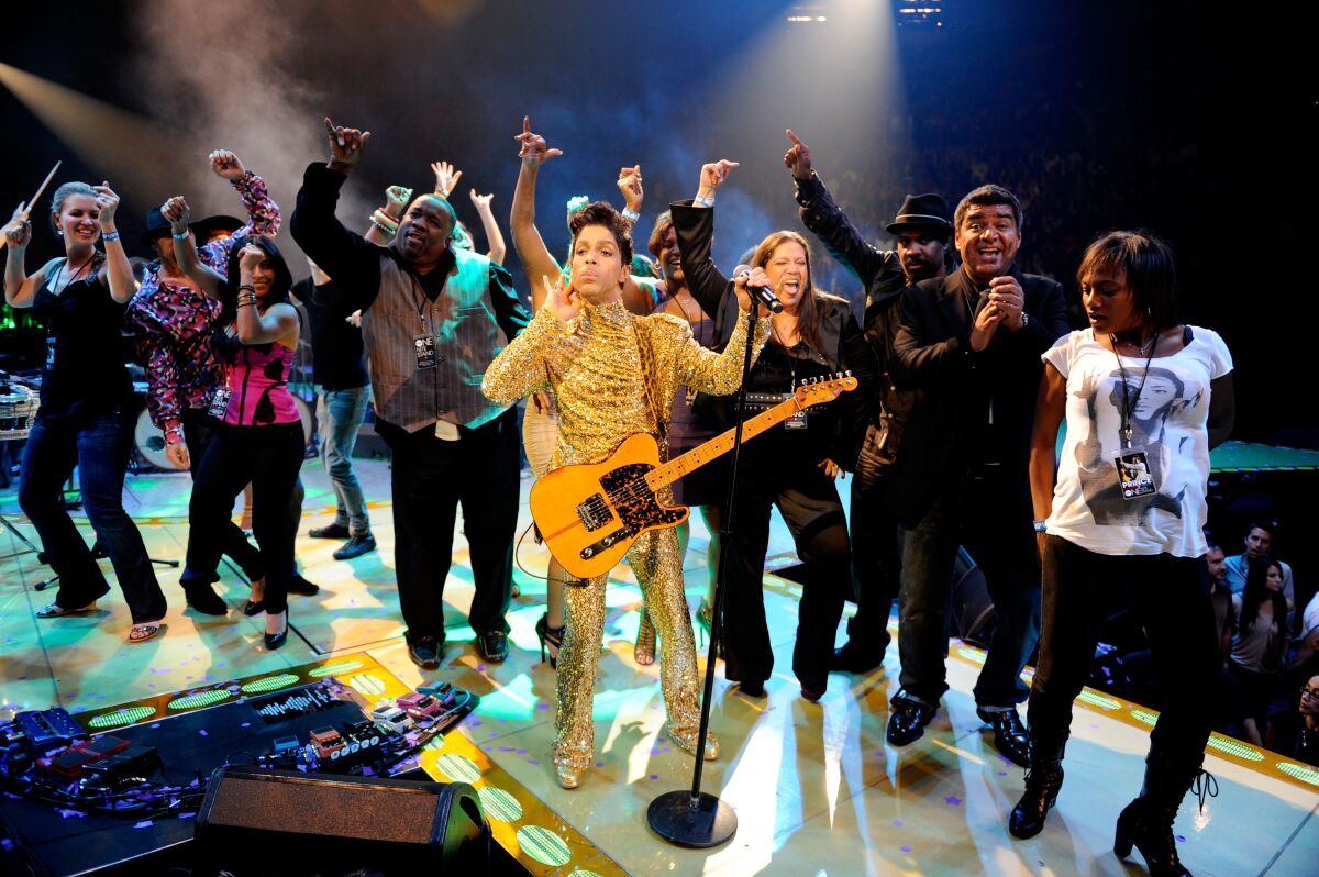 A man with a guitar is on a stage surrounded by background singers and musicians