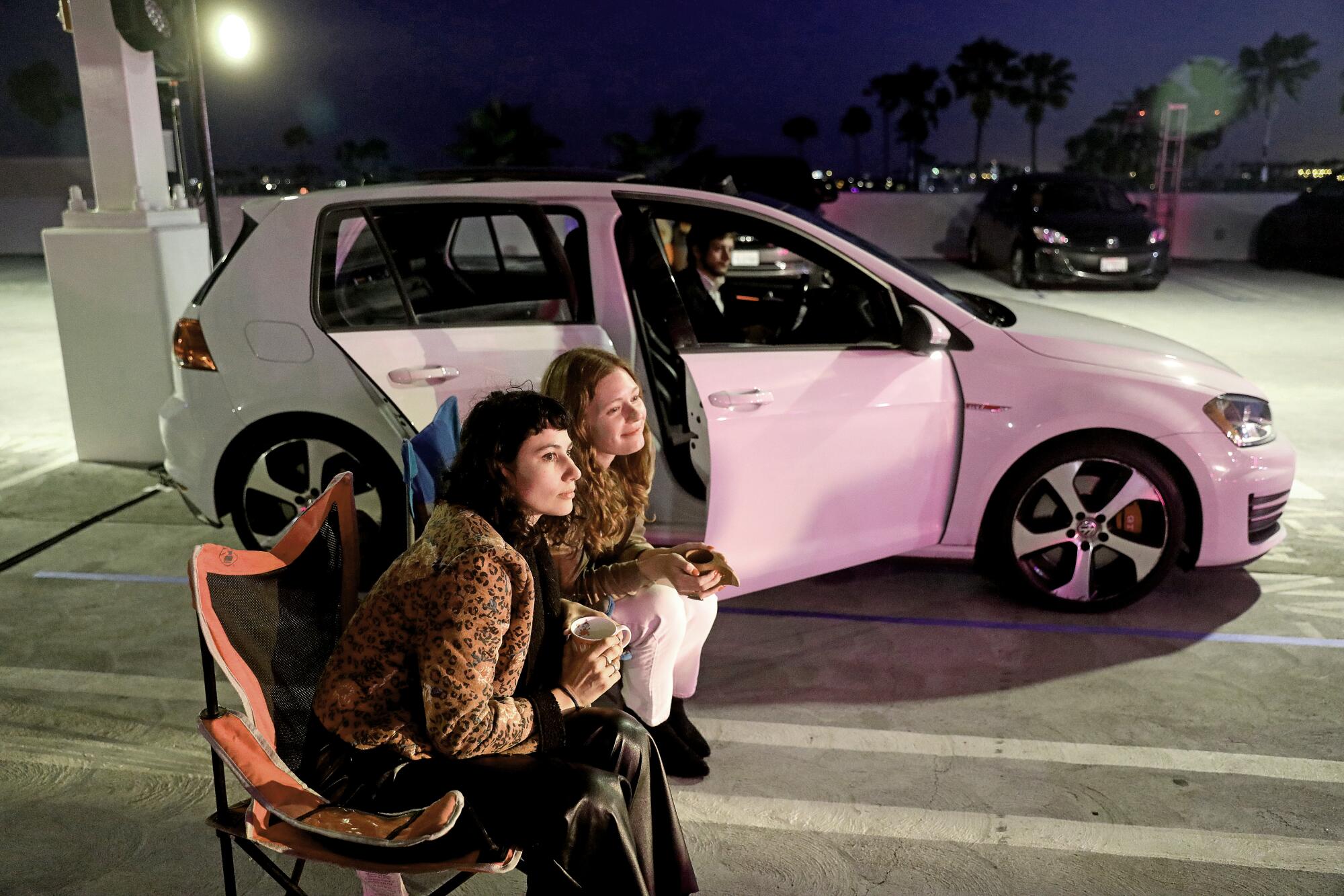 Two women sit in camp chairs beside a car with the passenger doors open with a man sitting in the driver's seat. 