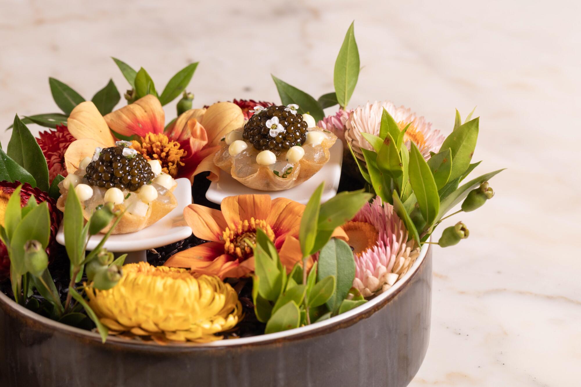 Small tarts topped with caviar and surrounded by flowers and greenery in a black bowl