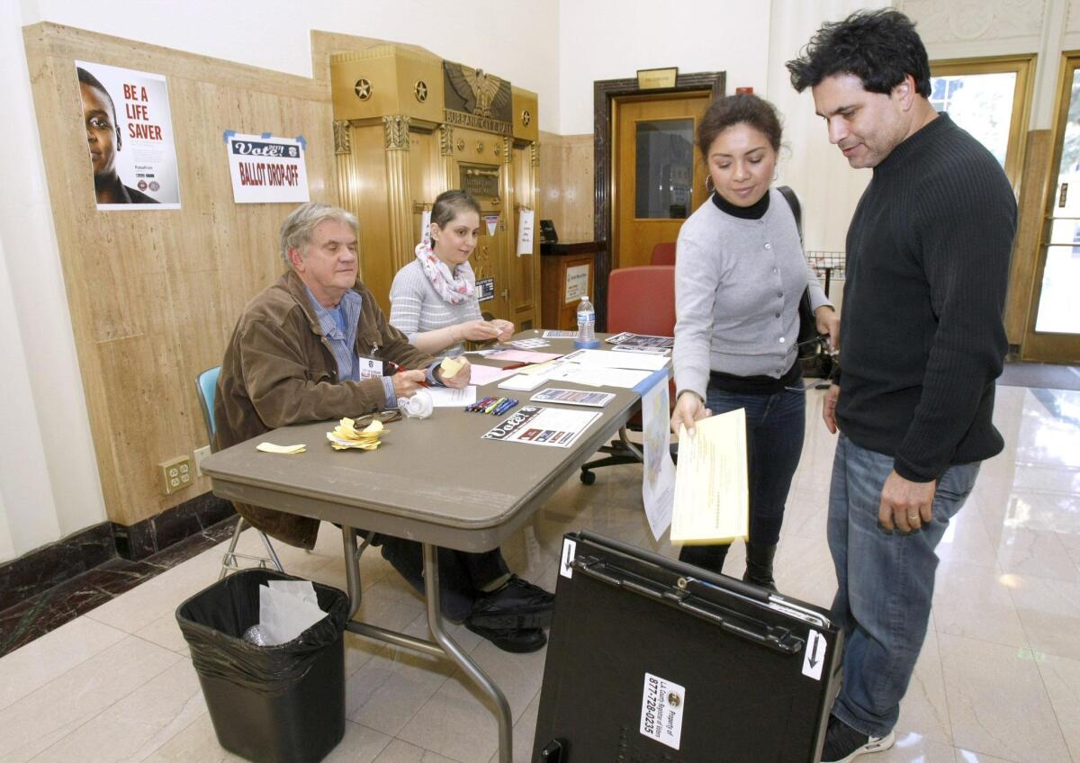 With poll center workers Kenneth Senior, left, and Angineh Goocherians looking on, Rosie Estrada and her husband Kevin Estrada drop off their ballots at city hall in Burbank on Tuesday.
