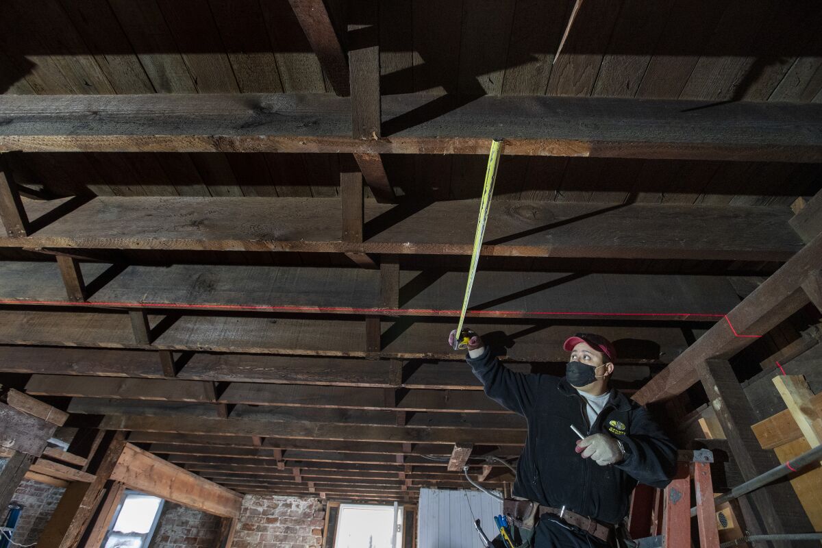 A worker on a ladder holds a tape measure to the boards of a ceiling.
