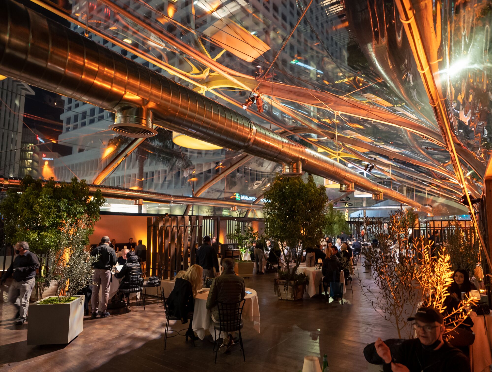 Diners eat inside a plastic modular at night, surrounded by trees and the skeleton of the structure