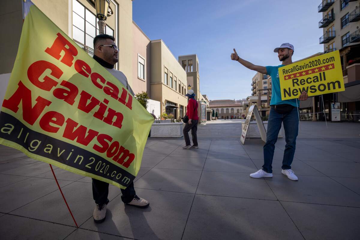 Volunteers hold up signs that say Recall Newsom and Recall Gavin Newsom outdoors at a shopping center.