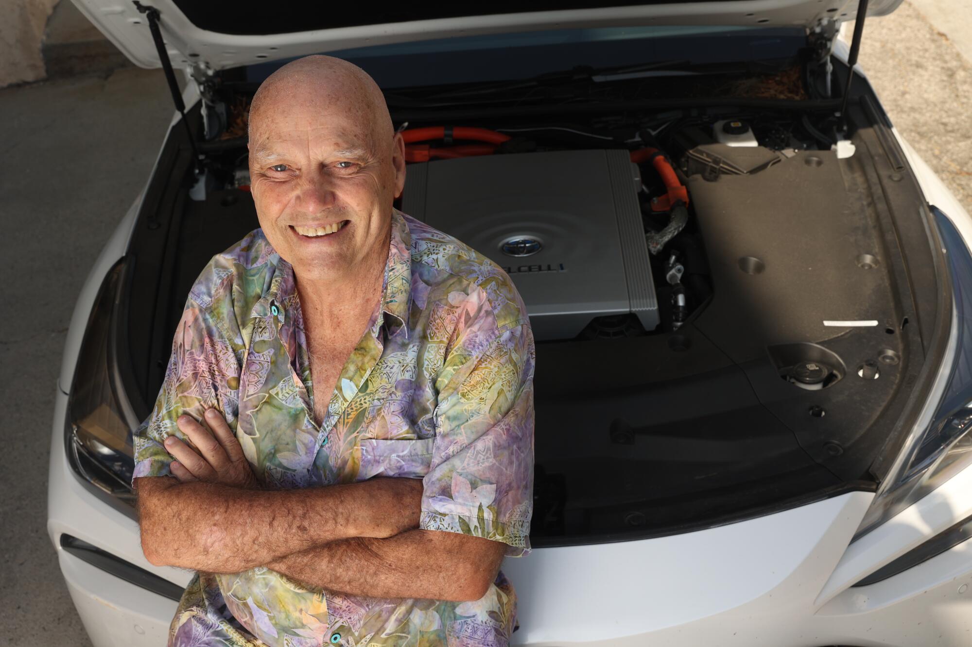 A man with bald head and flower patterned shirt crosses his arms with the hood of his car open behind him.