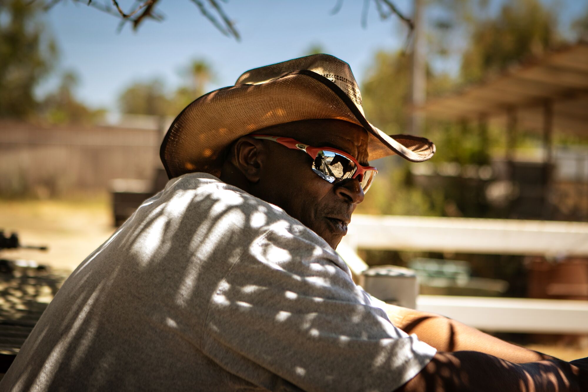 A close shot of a man wearing sunglasses and a cowboy hat, leaning on a fence 