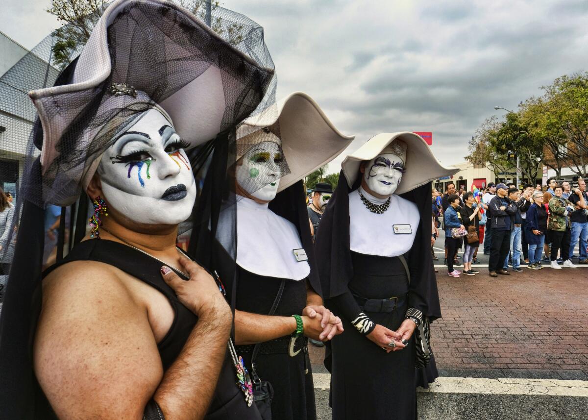 Three people in nun drag costumes stand on a street lined with onlookers. 