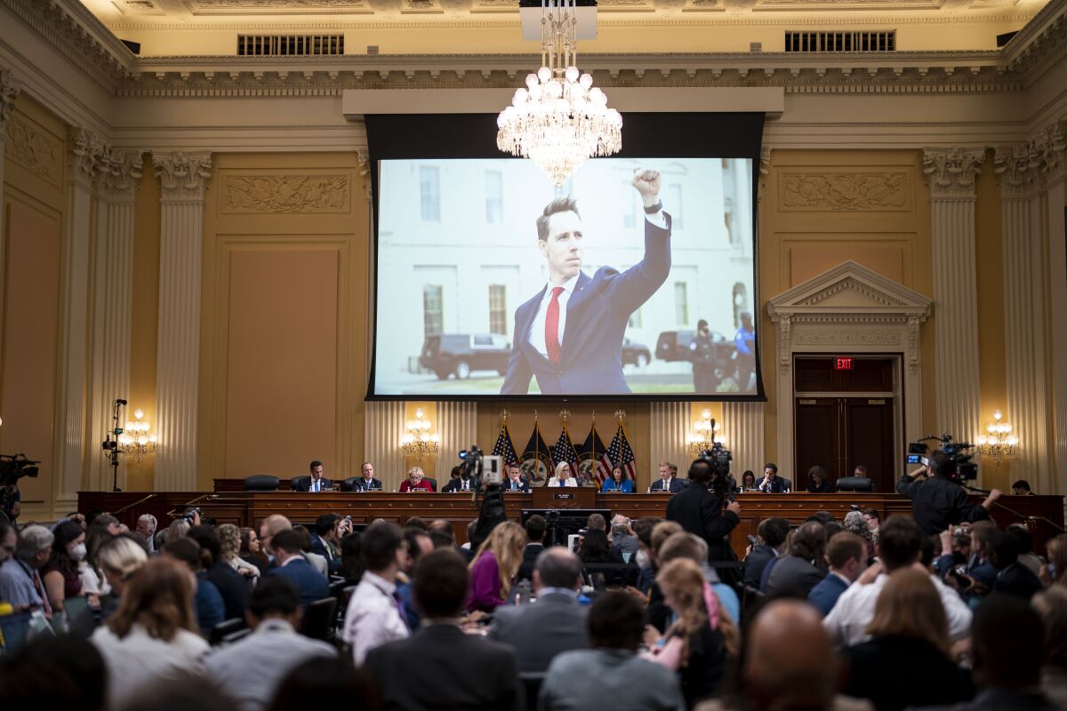 A large screen in a room full of people shows a man in a dark suit and red tie raising his hand, fist closed