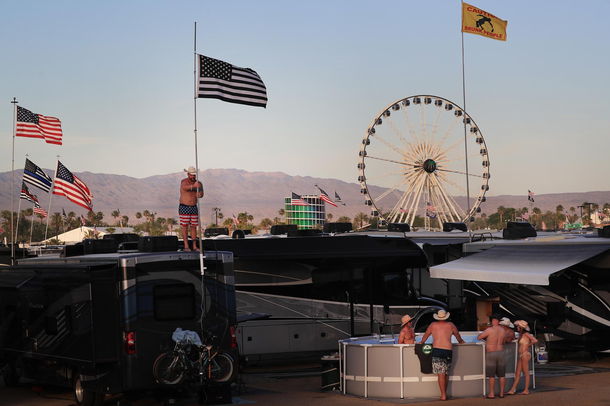 A man stands atop an RV raising an American flag as friends cool off in a portable pool with a Ferris wheel in the distance