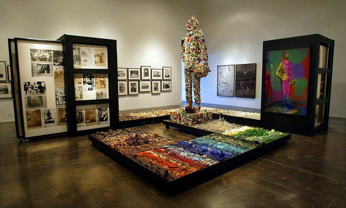 An installation view of Kelley's 2002 exhibition "Black Out" at Patrick Painter features the sculpture "John Glenn Memorial Detroit River Reclamation Project." It includes two big storage racks of archival newspaper stories connected by a low, trash-strewn platform. In the center is a larger-than-life statue of astronaut John Glenn, made from broken crockery.
