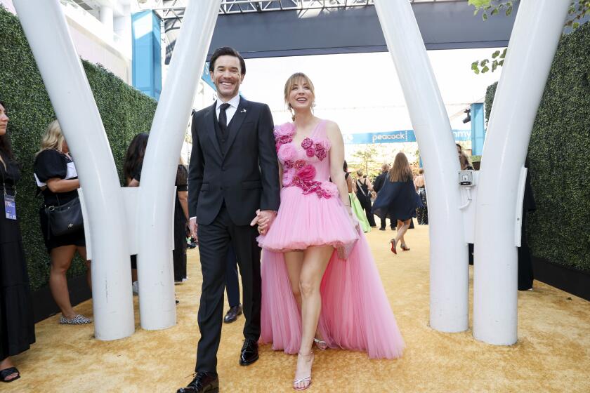 A man with dark hair in a black suit holding hands and walking with Kaley Cuoco who wears a pink tulle dress