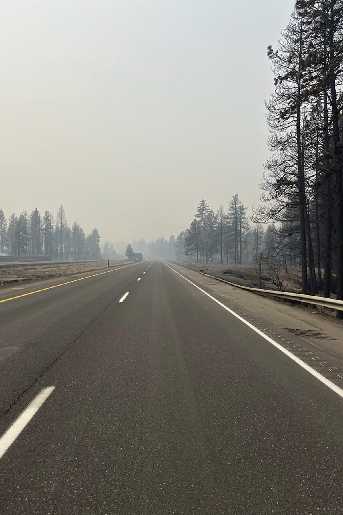 Smoke obscures blackened evergreens and the sky at the far end of a highway that stretches into the distance