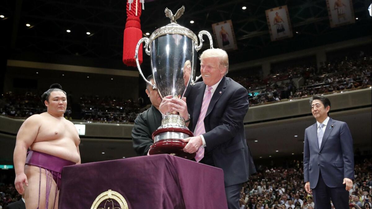 An official helps President Trump lift the President's Cup trophy to present it to Asanoyama, the Tokyo Grand Sumo Tournament winner, at Ryogoku Kokugikan Stadium on Sunday. Japanese Prime Minister Shinzo Abe is at right.
