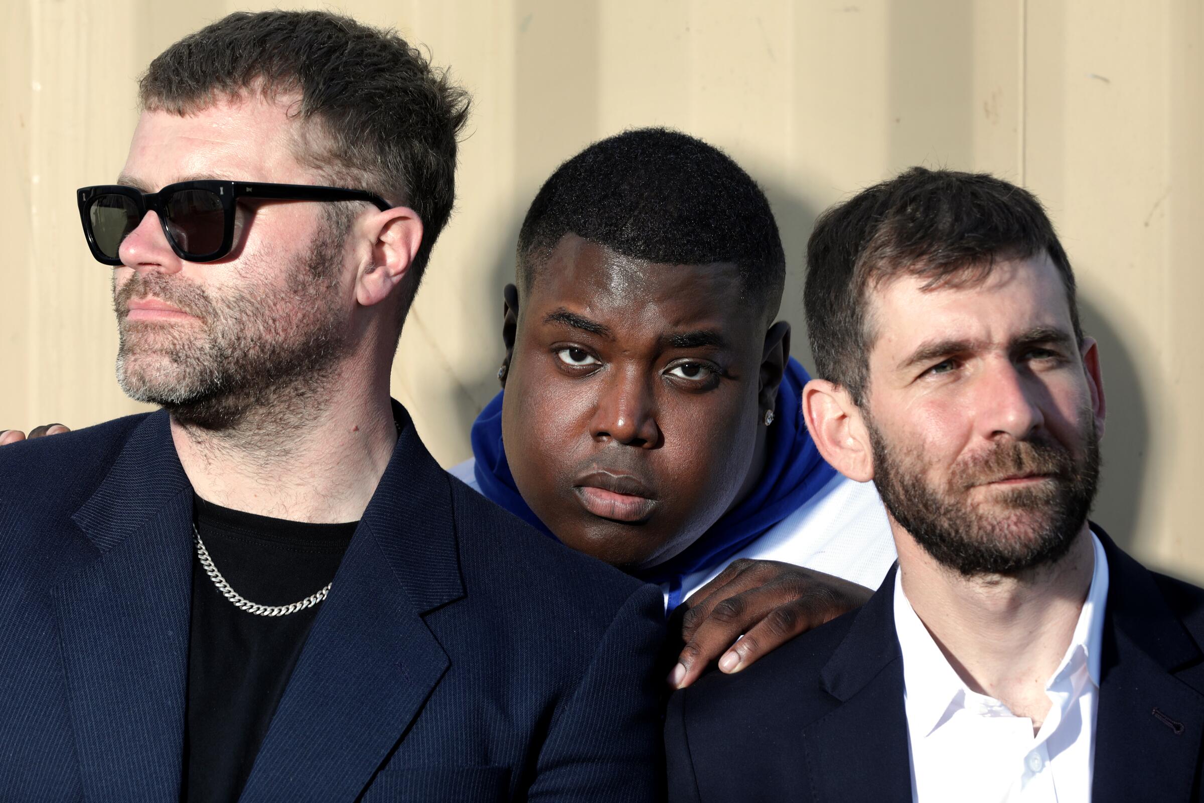 Three men, one wearing sunglasses, pose against a wall for a photo