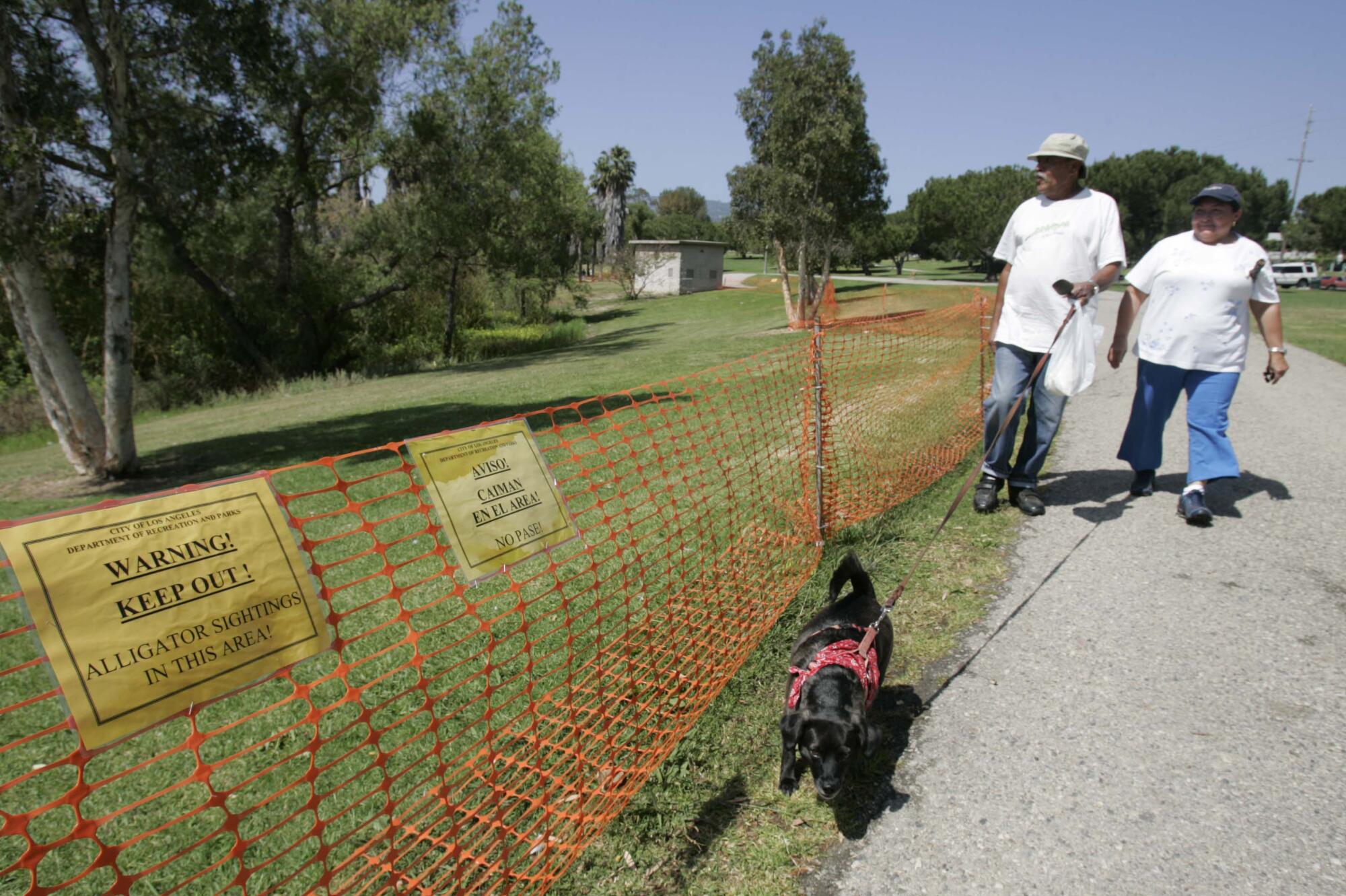 Two people walk a dog near fencing marked with with alligator warnings 