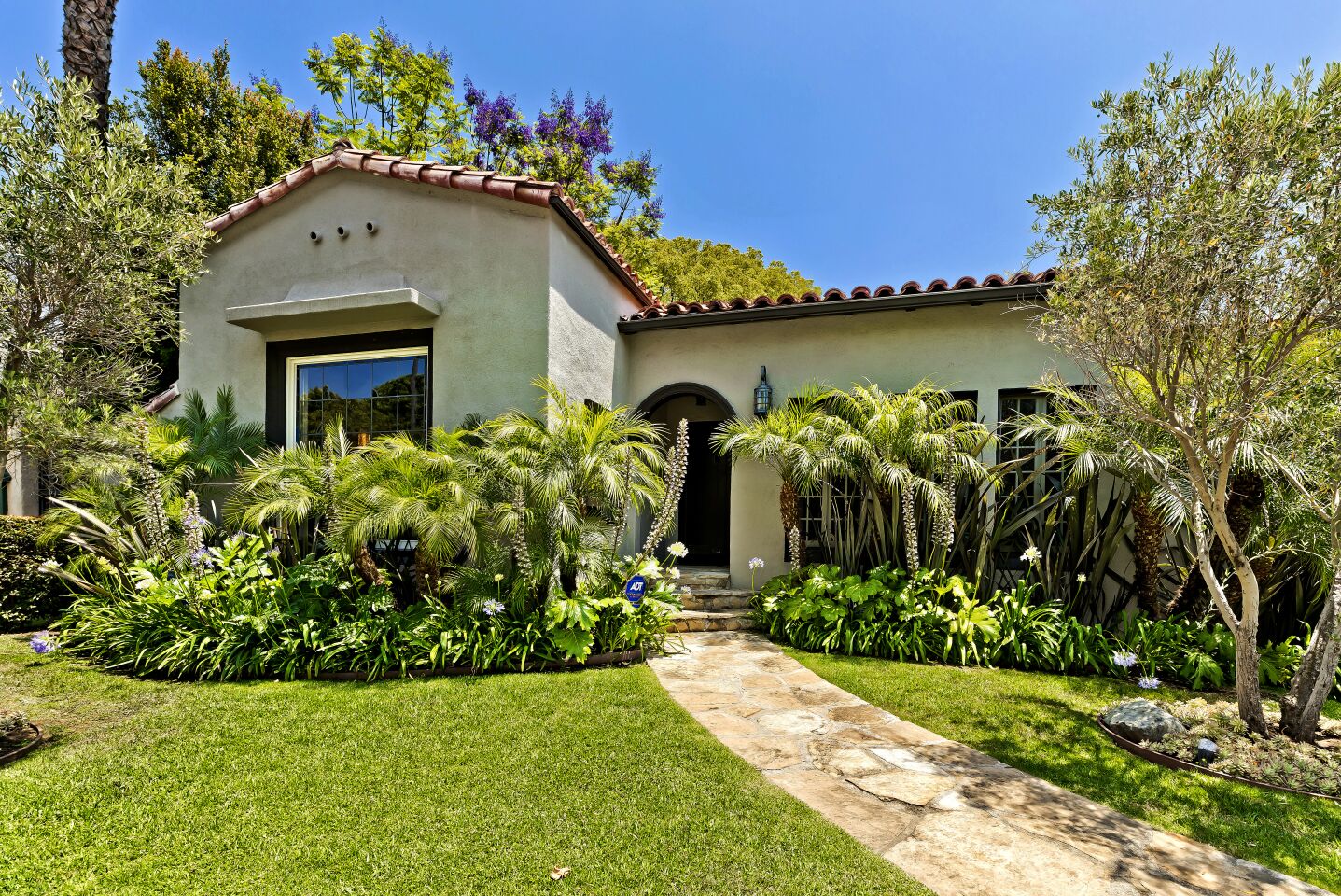 Exterior of a Spanish Colonial Revival style home with small palm trees, lawn and stone walkway