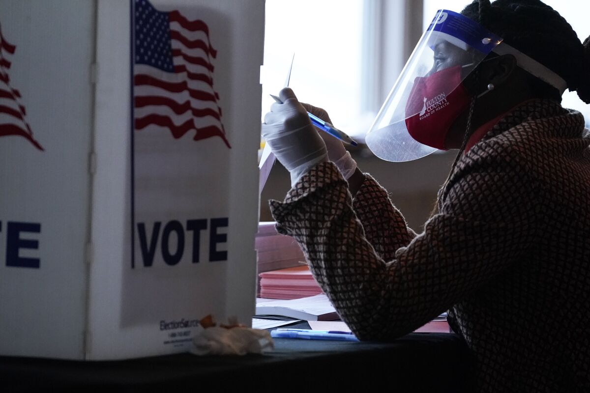 A poll worker wearing a face shield holds a ballot while sitting beside a ballot box 