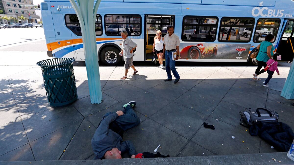 A homeless man sleeps under bus shelter while people exit an Orange County Transit Authority bus at Harbor Boulevard and Katella Avenue in Anaheim.