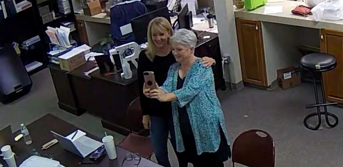 Two women smile toward a phone one is holding up in the middle of an elections office. 