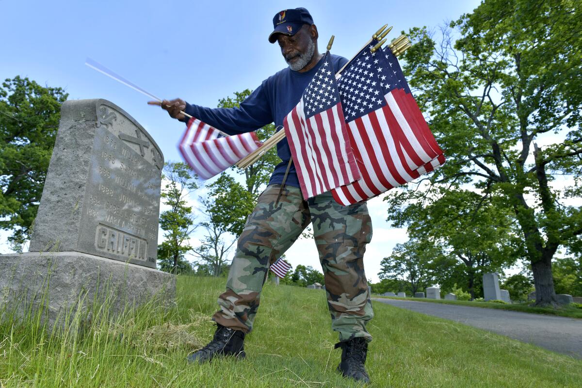 Bob Workman, holding small U.S. flags, stands alongside a gravestone.